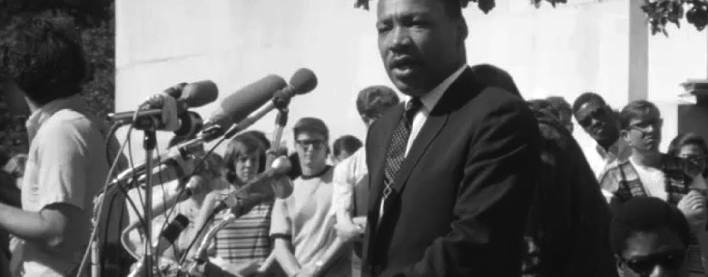 Dr. Martin Luther King Jr. speaks into a collection of microphones in front of a mostly white audience in a black-and-white still