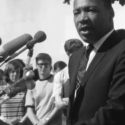 Dr. Martin Luther King Jr. speaks into a collection of microphones in front of a mostly white audience in a black-and-white still